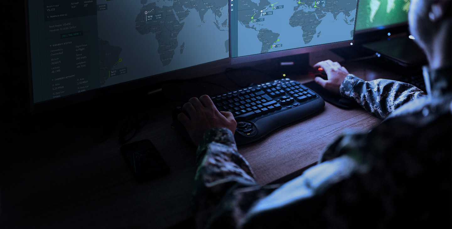 Close-up of a man dressed in a camo uniform working at a computer, looking at two monitors showing a map of the world