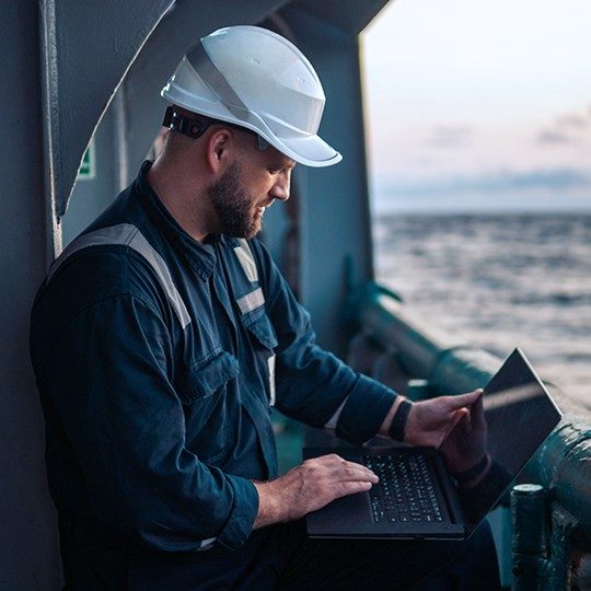 Man in a hard hat on a vessle at sea using maritime internet on a laptop.