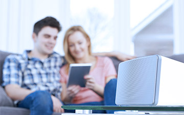 A connected smart speaker sits on a table in front of a  woman and man sitting on the couch looking at a tablet