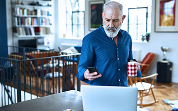 A man stands in front of a laptop at his table holding a coffee cup, using his smartphone