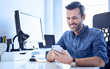 A man has home cyber security and sits in his home office on his smartphone smiling.