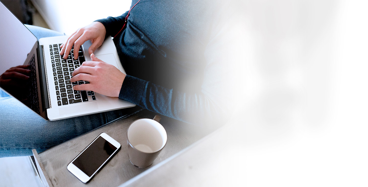 Close-up of a man casually sitting on a couch typing on his laptop with his phone and coffee cup next to him