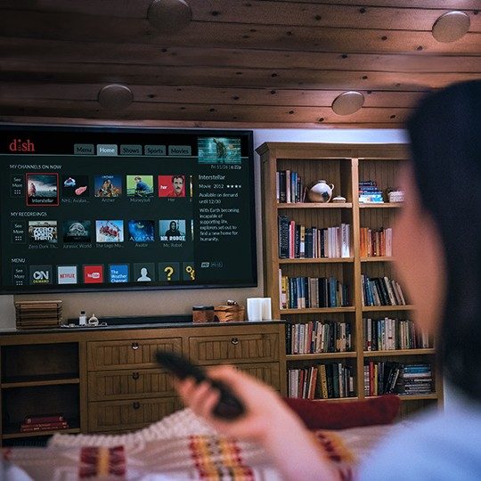 A woman streams content with her DISH TV and internet package, holding a remote and sitting on her couch