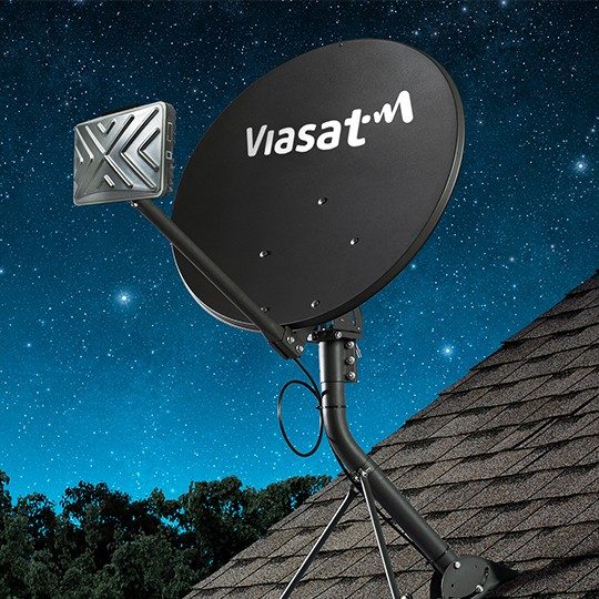 Gray fixed broadband terminal with TRIA and a white Viasat logo, mounted to the roof of a house against a starry night sky