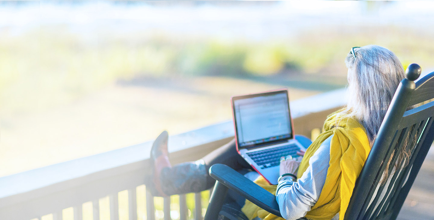 Woman in cowboy boots sitting in a rocking chair on her porch in teh countryside, typing on her laptop