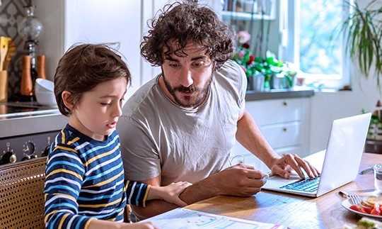 Man sits in front of his laptop at his kitched table, next to his son who is showing him sometimg on a paper