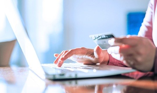 Close-up of a hand holding a card, typing into their laptop on a table