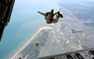 View from an open plane door of two military personnel skydiving towards a coastline
