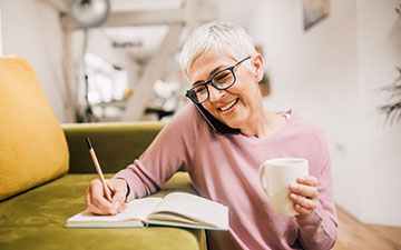 Woman sitting on the floor in front of a green couch, talking on the phone and writing in a book, holding a white cup