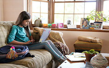 Young girl with long brown hair sitting on a couch next to her backpack doing homework on her laptop