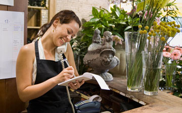 Woman working in a floral shop wearing a black apron, taking an order over the phone on a notepad