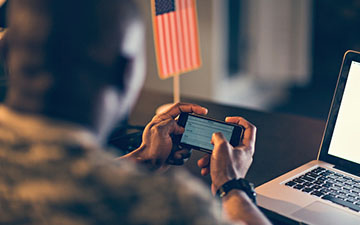 Close-up of a man in the military holding a smartphone horizontally in front of a laptop and american flag on a stand