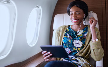 Professional woman with black hair sitting on a private plane, listening to content on a tablet with earbuds