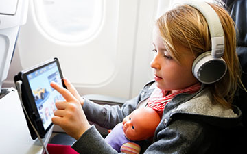 Young blonde girl sitting on a plane, holding a baby doll and wearing headphones, streaming content on a tablet