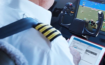 Pilot in uniform sitting in a plane's cockpit, holding a tablet showing Viasat document management software