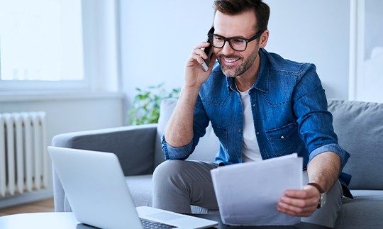 A man wearing glasses and a denim shirt talks on the phone while looking at his laptop