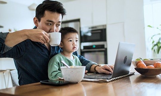 A man with a home phone and internet bundle sits at a table in his kitchen with his son on his lap, looking at a laptop