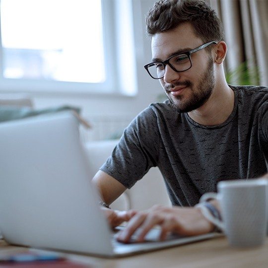 Man sitting at a table with laptop and cup of coffee staying connected with Viasat's satellite internet and the FCC's Affordable Connectivity Program.