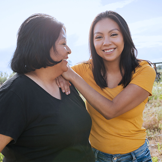 Two women smiling and standing shoulder to shoulder in an open field, excited to have affordable satellite internet with help from Viasat and the FCC's Affordable Connectivity Program.