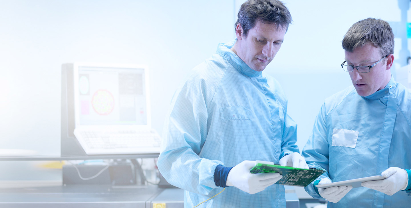 Two men in lab uniforms wearing latex gloves and looking at a semiconductor, one holding a tablet